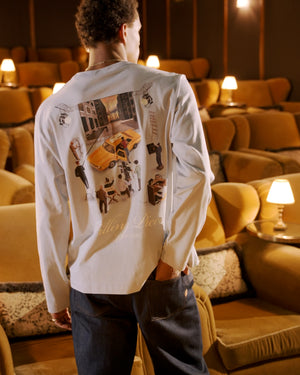 A man stands with his back to the camera in a dimly lit lounge, wearing a white long-sleeve shirt with a large multicolor graphic print on the back and dark jeans from filling pieces.